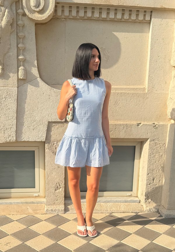 Femme aux cheveux foncés mi-longs portant une robe bleue à carreaux sans manches, des sandales blanches et tenant un petit sac à bandoulière.