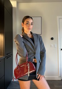 Grigia giacca su misura con spalle strutturate, abbinata a un top e una gonna neri. Tiene una borsa lucida color bordeaux con dettagli in catena.