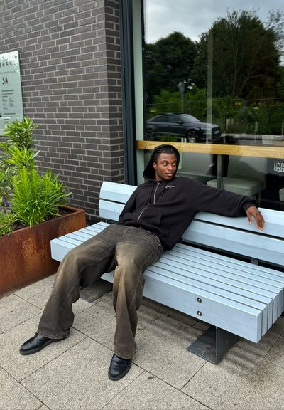 Banco de madera azul claro con diseño de listones. Una persona con una sudadera negra y pantalones marrones se sienta de manera casual contra una pared de ladrillo. Plantas verdes cercanas.