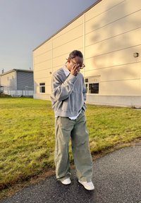 Overdimensjonert grå genser og vide olivengrønne bukser, kombinert med hvite joggesko. Står på gress ved siden av en bygning.