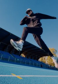 Crna flis jakna, crne hlače, bijele tenisice sa sivim potplatom i bijela kapa. Figura je u sredini skoka na plavoj stazi.