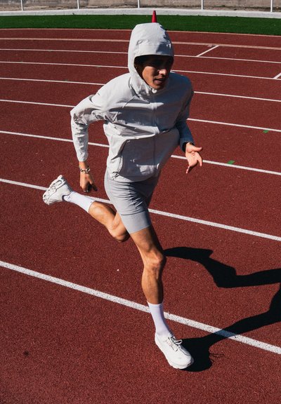 Corredor con sudadera gris con capucha y pantalones cortos corriendo a toda velocidad en una pista roja con marcas blancas en un estadio al aire libre soleado.