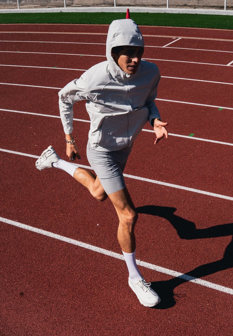 Løper i grå hettegenser og shorts som spurter på rød bane med hvite linjemarkeringer i solfylt utendørs stadion.