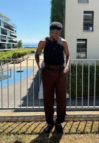 Svart ermeløs topp og mønstrede brune bukser med diamantformer. Svarte sko, stående på en balkong med grønt i bakgrunnen.