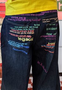 Gros plan sur un jean foncé avec des phrases colorées et dispersées comme "legacy", "honestly" et "for real" imprimées autour des poches arrière.