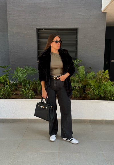 Abrigo de pelo sintético negro, blusa verde oliva, jeans negros de tiro alto, zapatillas blancas y un bolso negro. Los accesorios incluyen gafas de sol y pendientes.
