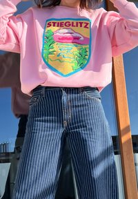 Sweatshirt rosa com um emblema bordado colorido apresentando imagens da natureza e o texto "STIEGLITZ." Acompanhado de jeans verticais listrados em azul escuro.