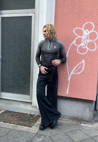 Tops de manga larga, ajustado, de color gris oscuro con un cuello con cremallera, combinado con pantalones negros de pierna ancha. Los accesorios incluyen múltiples anillos y pulseras.