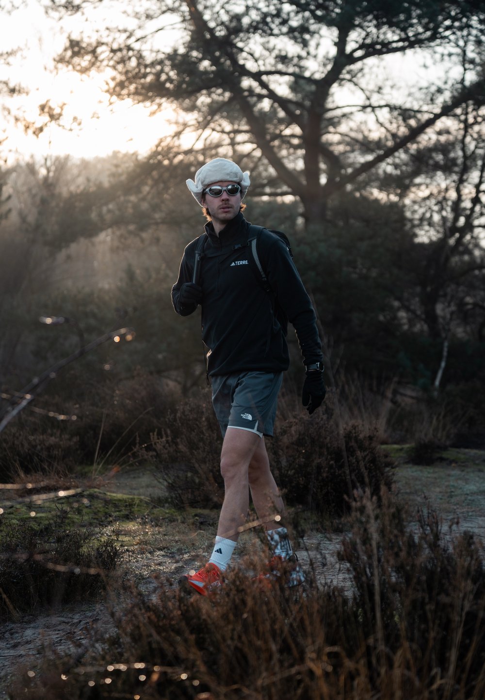 Un athlète portant une veste noire à fermeture éclair et un short gris, avec des gants blancs, des lunettes de soleil et des chaussures de course rouges, marche sur un chemin en plein air.