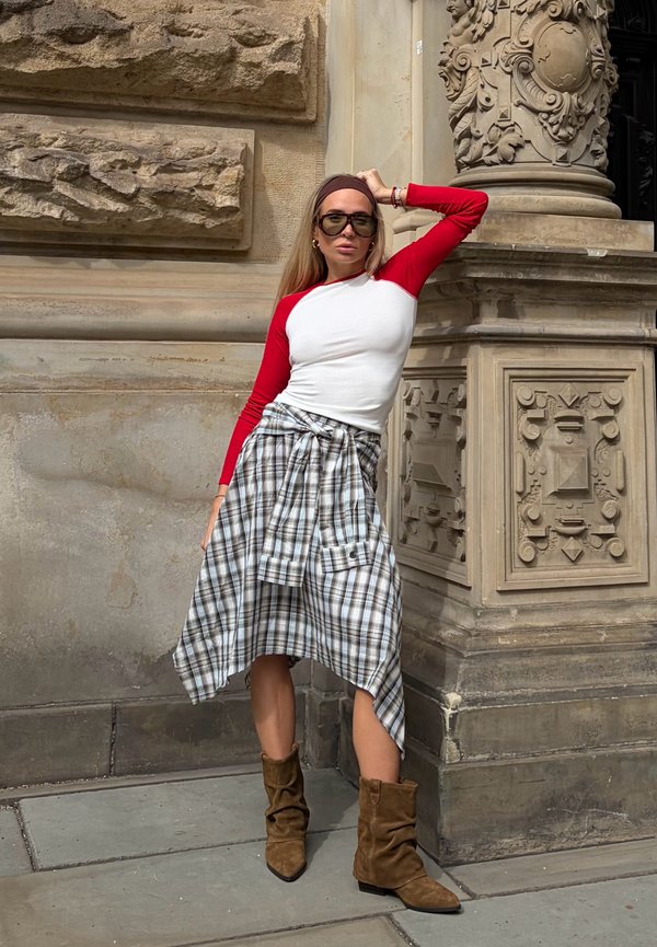 Femme aux longs cheveux portant des lunettes de soleil, une chemise rouge et blanche à manches longues, une jupe à carreaux et des bottes marron, appuyée contre un bâtiment en pierre orné.