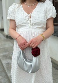 Kvinna i en vit texturerad klänning som håller en liten silverhandväska med en röd tygblomma, bär en guldklocka och ett pärlhalsband med bokstaven "A".
