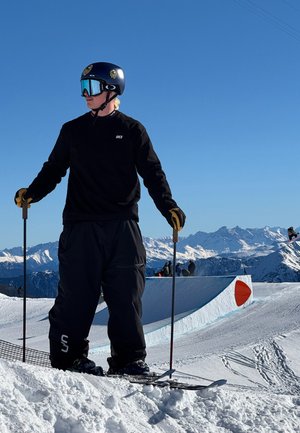 Equipo de esquí: camiseta negra de manga larga, pantalones anchos negros, casco azul, visor reflectante. De pie sobre los esquís con bastones, con un fondo de pendiente cubierta de nieve.