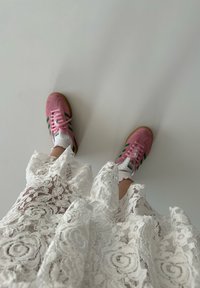 Sneakers rosa con strisce verdi, abbinati a un vestito bianco di pizzo con intricati motivi floreali e bordi arricciati.