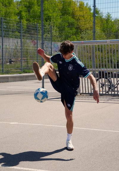 Joven con equipación deportiva azul marino hace malabares con una pelota de fútbol azul y blanca con la rodilla en una cancha exterior soleada, cerca de una valla metálica y árboles verdes.