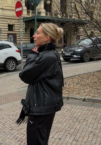 Jeune femme en veste en cuir noire et pantalon Puma se tient sur une rue pavée près de voitures et d'un panneau de circulation sens interdit.