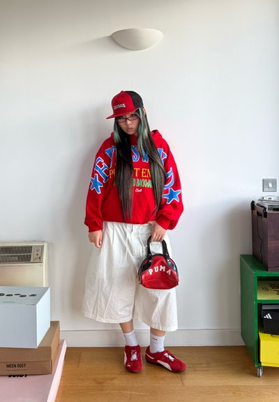 Joven con cabello largo que lleva una sudadera roja con capucha, pantalones anchos blancos, zapatillas rojas, una gorra negra con rojo y sostiene un bolso rojo de Puma en interiores.