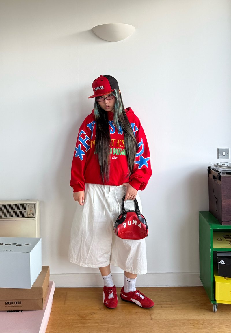 Jeune personne aux cheveux longs portant un sweat à capuche rouge, un pantalon blanc à jambes larges, des baskets rouges, une casquette noir-rouge, et tenant un sac à main Puma rouge à l'intérieur.