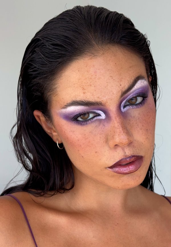Femme aux cheveux foncés mouillés, maquillage des yeux violet et blanc, lèvres violettes brillantes, petit anneau d'oreille, et haut à bretelles violet sur fond neutre.