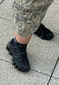 Mustad mustad Nike ketsid tekstuurse võrkpinnaga ja kummist aktsentidega, koos kamuflaažimustriga lõhikärgidega, asetatuna sillutatud pinnale.