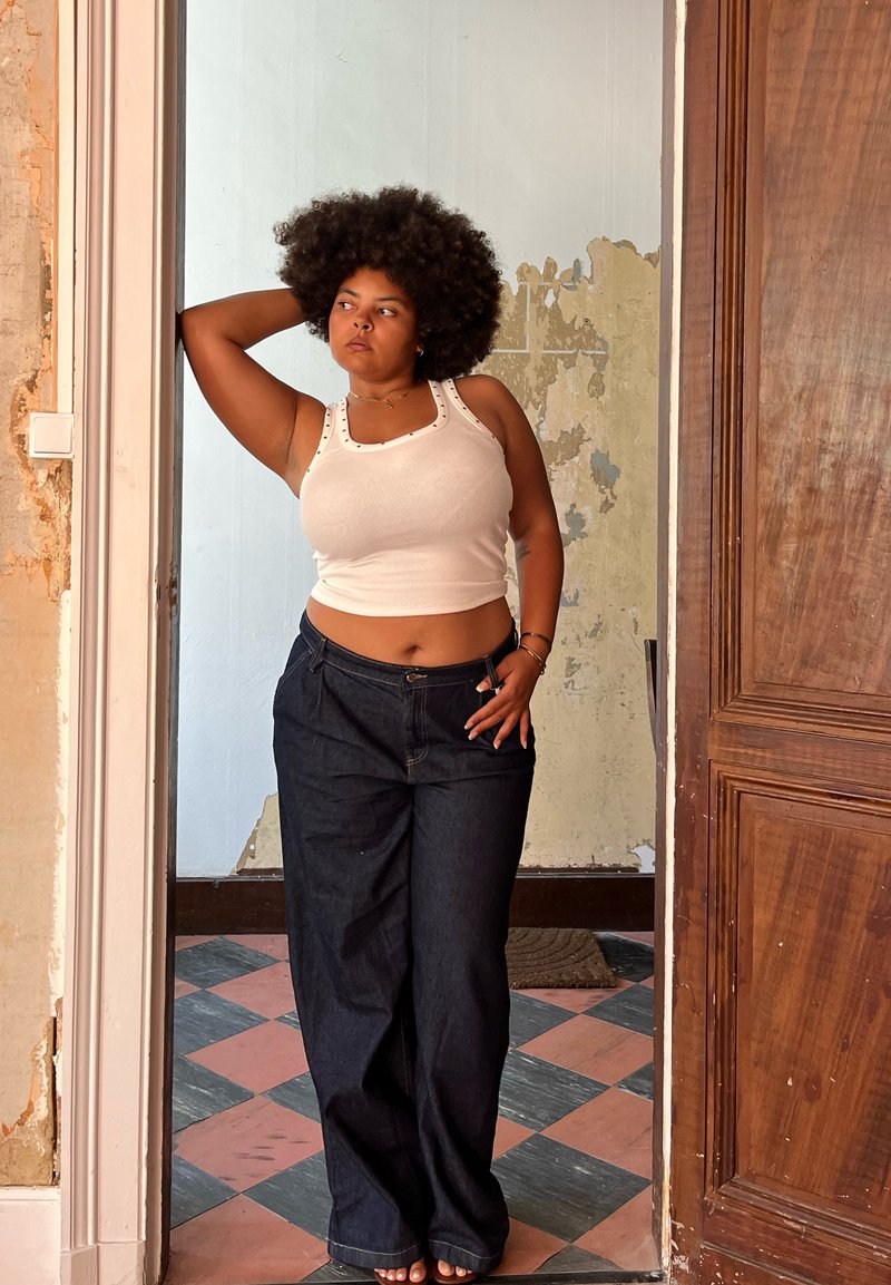 Camisola de alças branca com textura e jeans largos escuros, de pé numa porta. Paredes expostas e chão de azulejos decorados ao fundo.