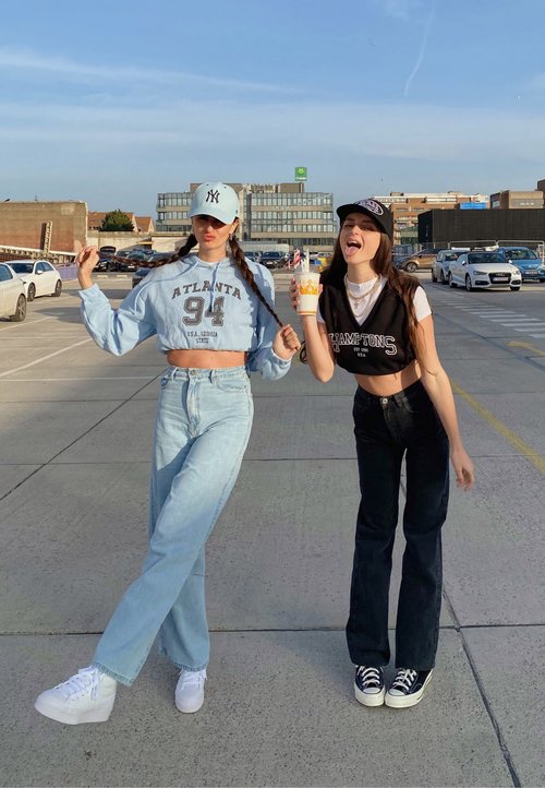 To kvinder står side om side. Den ene har en vest i pink og grøn argyle, mens den anden har en grøn cropped top med et "S." Begge iført lyse denim jeans og sneakers.