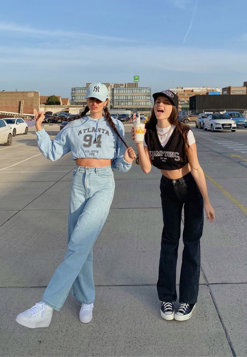 To unge kvinder står i et parkeringsområde. Den ene har en blå cropped sweatshirt, lyse jeans og hvide sneakers på. Den anden har en sort crop top, mørke jeans og sorte sneakers på.