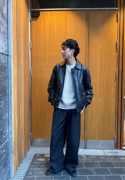 Chaqueta de cuero negra con grandes solapas, suéter de punto claro y pantalones negros holgados. De pie frente a una puerta de madera y una pared de piedra gris.