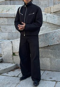 Black zip-up jacket with silver zipper accents, paired with loose-fitting black trousers and black shoes, standing on stone steps.