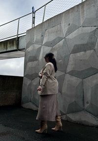 Beige blazer over en taupe plisseret nederdel, parret med nude højhælede støvler. Baggrunden er en struktureret grå betonvæg med geometriske mønstre.