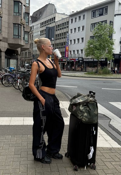 Top negro de tirantes y pantalones holgados negros con gráficos blancos, zapatillas negras. Lleva una bolsa negra, al lado de una maleta negra con ruedas y un bolso de lona de camuflaje.