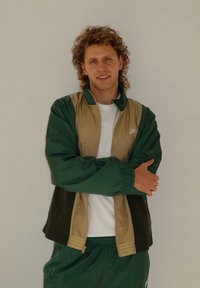 Noor mees lokkis juustega, seljas roheline ja beež Nike jakk ning rohelised püksid, seisab käed ristis lihtsa seina vastu.