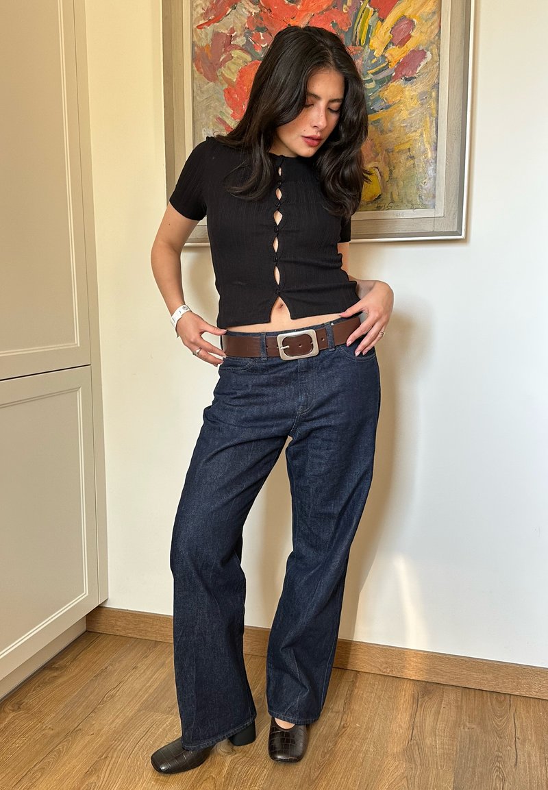 Femme aux cheveux foncés portant un haut noir à manches courtes boutonné, un jean foncé avec une ceinture marron et des chaussures noires, debout à l'intérieur.