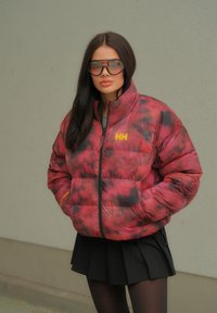 Κόκκινο και μαύρο πουπουλένιο μπουφάν με ψηλό γιακά, καρό σχέδιο και λογότυπο. Φοράει ένα μαύρο φούστα με λεπτομέρειες από κούμπωμα.