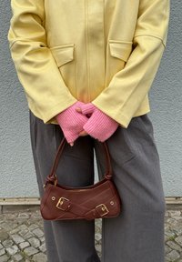 Borsa a tracolla in pelle marrone con hardware dorato, forma curva e tracolla, abbinata a una giacca giallo pallido e guanti senza dita rosa.