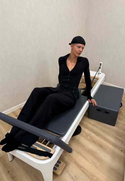 Máquina de Pilates Reformer con un tapete negro acolchado, barra de pie de madera y caja de resistencia gris. Usuario sentado con atuendo negro ajustado.