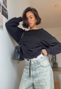 Jeune femme aux cheveux courts et foncés, portant un sweat-shirt noir oversize, un jean en denim clair et un sac à bandoulière, posant à l'intérieur près d'un mur blanc décoré.