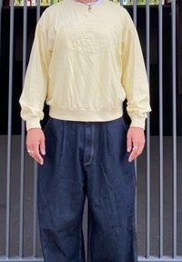 Sweatshirt amarelo com logótipo em relevo, com punhos canelados, combinado com calças de ganga escuras de corte largo, apresentando costuras visíveis. Fundo simples.