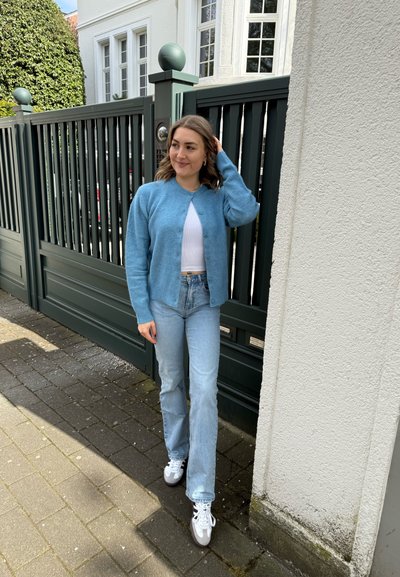 Cardigan azul claro, top blanco y jeans azul claro. Zapatillas blancas con acentos grises. De pie contra una puerta de madera verde.