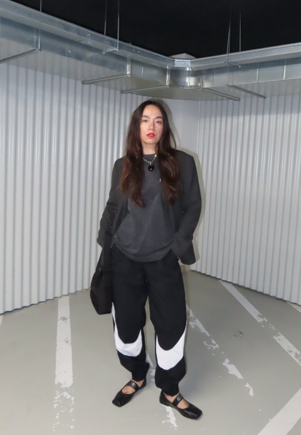 Femme aux longs cheveux noirs portant un pantalon noir et blanc, une veste sombre et des chaussures noires, debout dans une zone de stationnement industrielle aux murs ondulés.