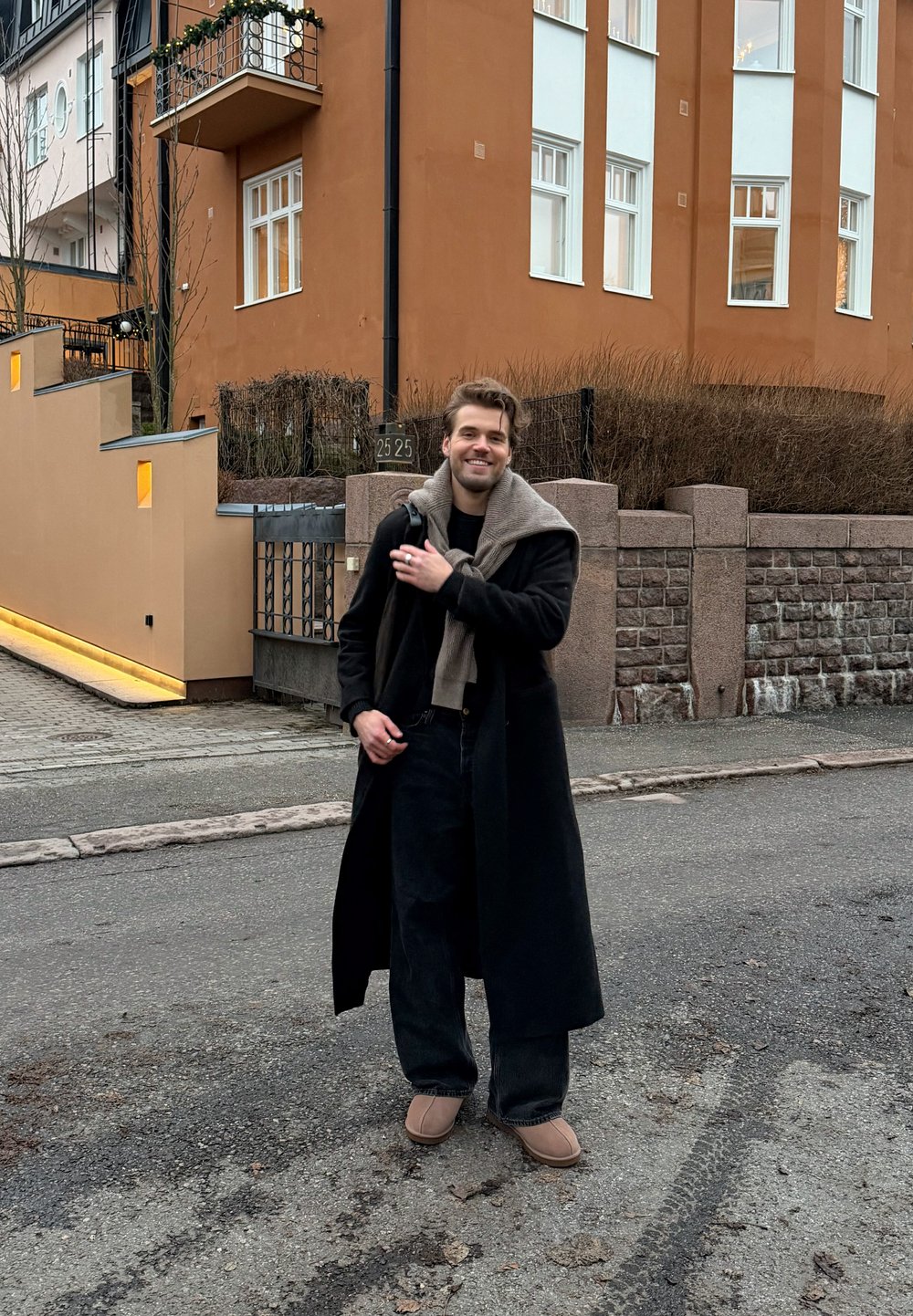 Manteau long noir, écharpe grise posée sur les épaules, pantalon noir et chaussures beiges. En arrière-plan, un bâtiment contemporain et un chemin.