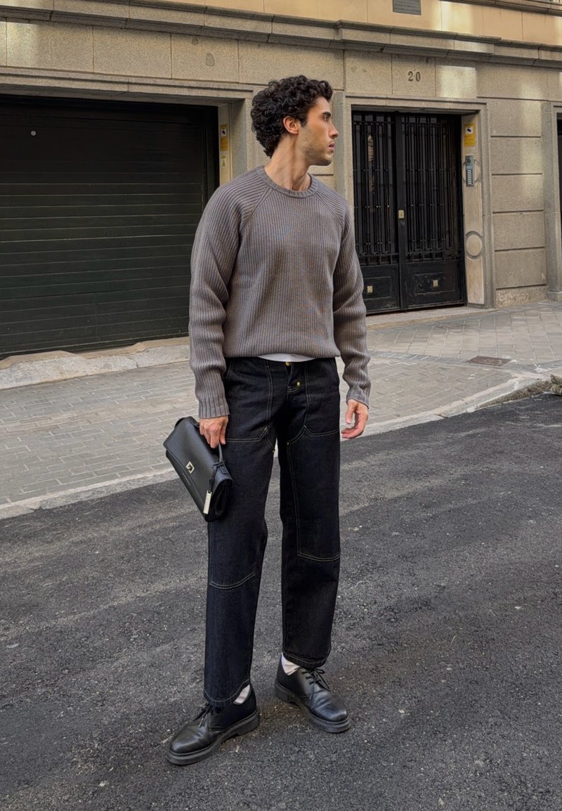 Jeune homme portant un pull gris, un jean noir et des chaussures noires, tenant une pochette noire, debout dans une rue de la ville, regardant vers la droite.