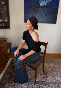 Femme aux cheveux foncés relevés en chignon, portant un haut noir épaules dénudées et un jean bleu, assise en tailleur sur une chaise en bois dans une pièce décorée.