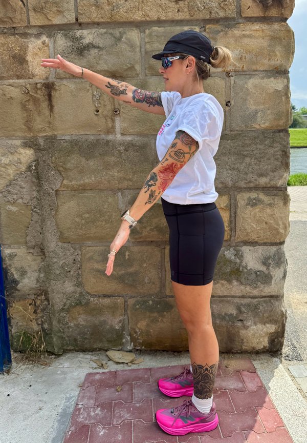Femme avec des tatouages en tenue de sport, debout de profil, les bras tendus parallèlement au sol, contre un mur en pierre en extérieur.