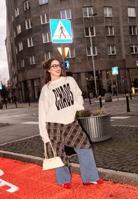 Pull surdimensionné gris clair avec le texte noir "CHAOS", jupe à carreaux superposée, jean évasé en denim, chaussures rouges et sac à main crème dans un décor urbain.