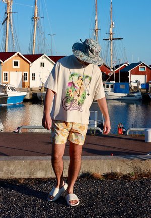 Camiseta de manga corta de color crema con gráfico floral y texto "Pivitti", combinada con pantalones cortos amarillos con flores y sandalias blancas de punta abierta.