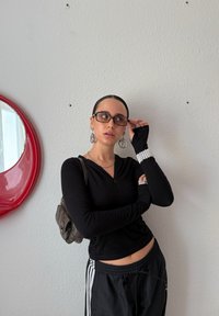 Cămașă neagră cu mânecă lungă și fermoar, pantaloni negri cu dungi albe, cercei rotunzi din argint și ochelari. Oglindă circulară roșie montată pe perete în fundal.