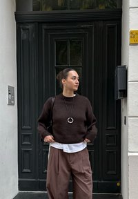 Frau mit dunklem Haar im Dutt, die einen dunkelbraunen Pullover, ein weißes Hemd und lockere braune Hosen trägt, steht vor einer schwarzen Tür mit den Händen in den Taschen.