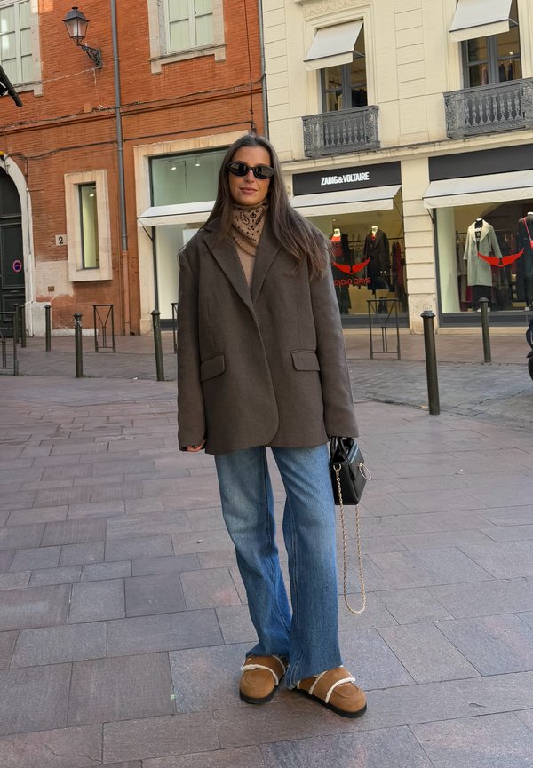 Brun oversized blazer, beige striktrøje, flagrende lyse denim jeans, brune slippers med hvide detaljer, sort håndtaske med kædestrop.