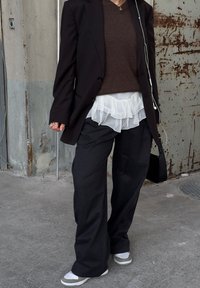 Persona, kas ģērbjas brūnā džemperī, melnā bleizerī, ar mīksto baltu blūzi, platām melnām biksēm un sneakers apaviem, tur melnu rokassomu blakus rūsējošai metāla sienai.