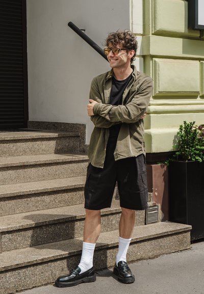Camisa de botones verde, camiseta negra, pantalones cortos de carga negros, calcetines blancos y mocasines negros, de pie en escalones de piedra junto a una planta.